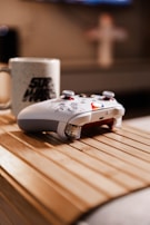 A white gaming controller with red accents rests on a wooden surface. Next to it, there is a white mug with a black logo printed on it. The background is slightly blurred, adding depth to the focus on the controller and the mug.
