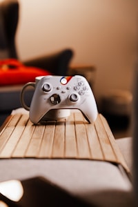 A white gaming controller with various buttons and joysticks is placed on a wooden surface. The controller has colored accents near the logo area. In the background, there is blurred furniture, adding a cozy indoor atmosphere.