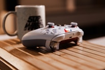 A gaming controller with a custom design rests on a wooden surface, next to a ceramic mug decorated with a recognizable logo. The lighting creates a warm and cozy ambiance, highlighting the textures of the materials.