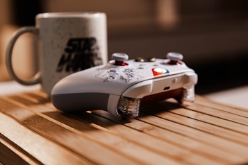 A gaming controller with a custom design rests on a wooden surface, next to a ceramic mug decorated with a recognizable logo. The lighting creates a warm and cozy ambiance, highlighting the textures of the materials.