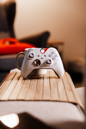 A white gaming controller rests on the rim of a white mug, placed on a wooden tray. The background is blurred, suggesting a cozy room setup with soft lighting and a hint of a red cushion or object.