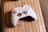 A white gaming controller with red accents and various buttons lies on a wooden surface with a light brown color and grooved texture. The controller features a gold-colored directional pad and buttons labeled with letters. The design includes colorful stripes on the top and small, precise markings on its surface.