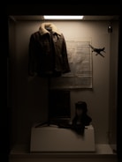 A museum or display case containing a leather jacket with a fur collar hanging on a mannequin. Below the jacket is a mannequin head wearing an aviator hat and goggles. In the background, there's a map on the wall, and a small model airplane is suspended in the air.