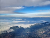 An aerial view over a landscape showing a vast expanse of clouds covering the land. Below the clouds, rugged terrain with mountains and valleys is visible. Above, the sky transitions from a vibrant blue to areas covered by thick, gray clouds.