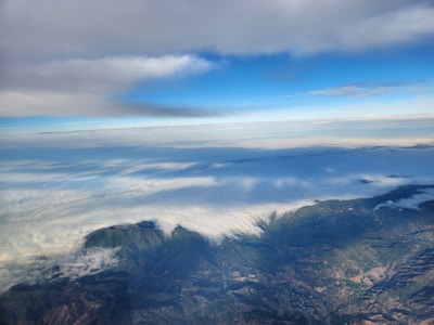 An aerial view over a landscape showing a vast expanse of clouds covering the land. Below the clouds, rugged terrain with mountains and valleys is visible. Above, the sky transitions from a vibrant blue to areas covered by thick, gray clouds.