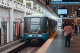 A modern train is stationed at an urban platform with passengers walking on the platform. The train has a sleek design with visible signage. Advertisements line the walls of the station, and overhead signs display departure information.
