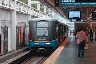A modern train is stationed at an urban platform with passengers walking on the platform. The train has a sleek design with visible signage. Advertisements line the walls of the station, and overhead signs display departure information.