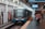 A modern train is stationed at an urban platform with passengers walking on the platform. The train has a sleek design with visible signage. Advertisements line the walls of the station, and overhead signs display departure information.
