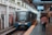 A modern train is stationed at an urban platform with passengers walking on the platform. The train has a sleek design with visible signage. Advertisements line the walls of the station, and overhead signs display departure information.