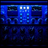 A close-up view of an audio processing panel, featuring various knobs, dials, and buttons. The panel displays intricate details such as compression meters, EQ controls, and frequency adjustments. The lighting casts a deep blue hue over the entire panel, giving it a high-tech, sophisticated appearance.