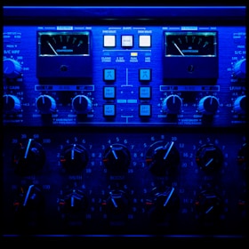 A close-up view of an audio processing panel, featuring various knobs, dials, and buttons. The panel displays intricate details such as compression meters, EQ controls, and frequency adjustments. The lighting casts a deep blue hue over the entire panel, giving it a high-tech, sophisticated appearance.