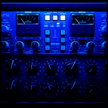 A close-up view of an audio processing panel, featuring various knobs, dials, and buttons. The panel displays intricate details such as compression meters, EQ controls, and frequency adjustments. The lighting casts a deep blue hue over the entire panel, giving it a high-tech, sophisticated appearance.