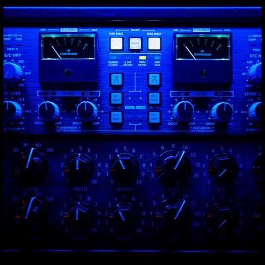A close-up view of an audio processing panel, featuring various knobs, dials, and buttons. The panel displays intricate details such as compression meters, EQ controls, and frequency adjustments. The lighting casts a deep blue hue over the entire panel, giving it a high-tech, sophisticated appearance.