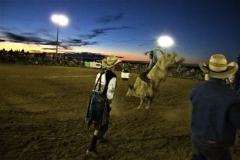 In a rodeo arena during sunset, a cowboy rides a bucking bull while two other cowboys, both wearing hats, observe nearby. The arena is dimly lit by bright floodlights, and a crowd watches from the stands in the background.