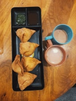 Four fried pastries are arranged on a rectangular black tray accompanied by two small bowls of green and brown chutneys. Beside the tray, there are two ceramic mugs filled with tea, one blue and the other orange. The setup is placed on a wooden table.
