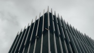 A modern building with a striking architectural design. Vertical metal or concrete fins protrude from the facade, creating an angular and geometric aesthetic. The overcast sky adds a moody and subdued background to the sharp, clean lines of the structure.