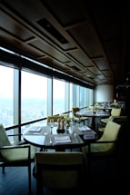 A modern, upscale restaurant with large floor-to-ceiling windows offering a cityscape view. Round tables with neatly arranged place settings and comfortable upholstered chairs fill the dining area. The ceiling features a geometric wooden panel design, creating a sophisticated atmosphere. The soft lighting enhances the elegant ambiance.