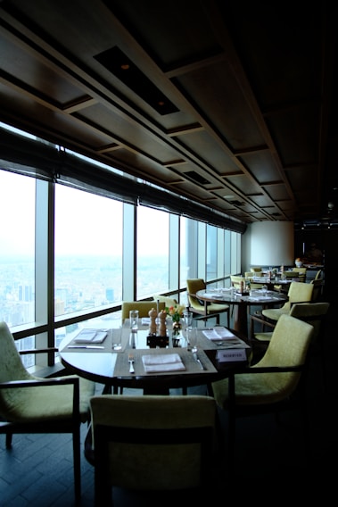 A modern, upscale restaurant with large floor-to-ceiling windows offering a cityscape view. Round tables with neatly arranged place settings and comfortable upholstered chairs fill the dining area. The ceiling features a geometric wooden panel design, creating a sophisticated atmosphere. The soft lighting enhances the elegant ambiance.