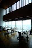 A modern, upscale dining area with wooden floors and large panoramic windows providing a view of a cityscape. The tables are neatly arranged with chairs, and there are small floral centerpieces on each table. The high ceiling and ambient lighting give the room an elegant feel.