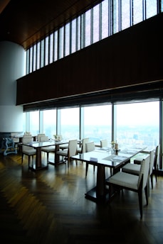 A modern, upscale dining area with wooden floors and large panoramic windows providing a view of a cityscape. The tables are neatly arranged with chairs, and there are small floral centerpieces on each table. The high ceiling and ambient lighting give the room an elegant feel.