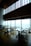A modern, upscale dining area with wooden floors and large panoramic windows providing a view of a cityscape. The tables are neatly arranged with chairs, and there are small floral centerpieces on each table. The high ceiling and ambient lighting give the room an elegant feel.