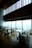 A modern, upscale dining area with wooden floors and large panoramic windows providing a view of a cityscape. The tables are neatly arranged with chairs, and there are small floral centerpieces on each table. The high ceiling and ambient lighting give the room an elegant feel.