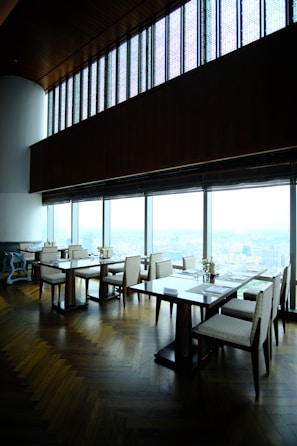 A modern, upscale dining area with wooden floors and large panoramic windows providing a view of a cityscape. The tables are neatly arranged with chairs, and there are small floral centerpieces on each table. The high ceiling and ambient lighting give the room an elegant feel.