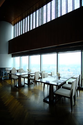 A modern, upscale dining area with wooden floors and large panoramic windows providing a view of a cityscape. The tables are neatly arranged with chairs, and there are small floral centerpieces on each table. The high ceiling and ambient lighting give the room an elegant feel.