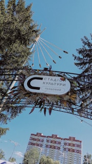 A decorative archway adorned with leaves and a sign with Cyrillic text stands amidst tall pine trees. The sky is clear and blue, with a complex structure resembling a sun with rays extending upwards. Below the structure, a multicolored apartment building is visible in the background.