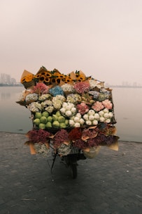 A motorbike is heavily laden with vibrant and diverse bouquets of flowers, including sunflowers and lilies. The bike is parked on a paved riverside with a calm body of water and a hazy cityscape in the background.