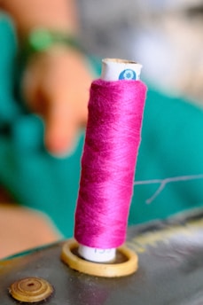 A spool of bright pink thread is mounted on a sewing machine. In the background, there is a blurred human hand and a green fabric, possibly indicating sewing activity.