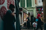 A narrow street scene featuring several people going about their daily activities. To the left, a person is standing against a wall decorated with red Chinese motifs, seemingly engaged in a phone call. In the center, a couple, both wearing hats, are seen with one person sitting in a wheelchair while the other stands beside. To the right, a man wearing a mask and dressed in a red shirt walks past the scene with a turquoise bag. The background includes shops with signage and upper floor building facades painted in green and white.