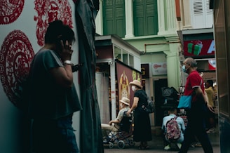 A narrow street scene featuring several people going about their daily activities. To the left, a person is standing against a wall decorated with red Chinese motifs, seemingly engaged in a phone call. In the center, a couple, both wearing hats, are seen with one person sitting in a wheelchair while the other stands beside. To the right, a man wearing a mask and dressed in a red shirt walks past the scene with a turquoise bag. The background includes shops with signage and upper floor building facades painted in green and white.