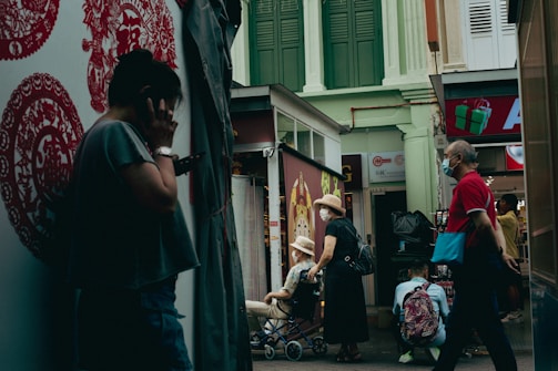 A narrow street scene featuring several people going about their daily activities. To the left, a person is standing against a wall decorated with red Chinese motifs, seemingly engaged in a phone call. In the center, a couple, both wearing hats, are seen with one person sitting in a wheelchair while the other stands beside. To the right, a man wearing a mask and dressed in a red shirt walks past the scene with a turquoise bag. The background includes shops with signage and upper floor building facades painted in green and white.