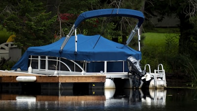 A blue canopied pontoon boat is docked on a wooden pier surrounded by green trees and foliage. The boat has metal railings and an outboard motor attached at the back. The water reflects the boat and the surrounding greenery, creating a serene waterfront scene.
