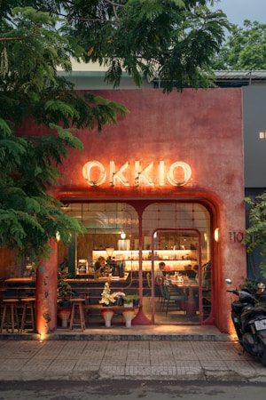 A quaint caf&eacute; with a rustic, earthy exterior features a prominent, illuminated sign reading 'OKKIO'. The fa&ccedil;ade is adorned with warm, reddish-brown tones and framed by lush green trees. Large glass windows provide a glimpse into the cozy and inviting interior where people are seated and engaged in conversation. Potted plants and wooden stools are positioned outside, enhancing the charming ambiance. A motorbike is parked near the entrance on the right side.