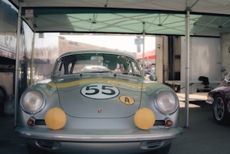 A vintage silver race car is parked under a canopy. The car features a racing number 55 on the hood and two yellow accents on the headlights. It is positioned in a setting typical for a motorsport event, with additional vehicles and equipment visible in the background.