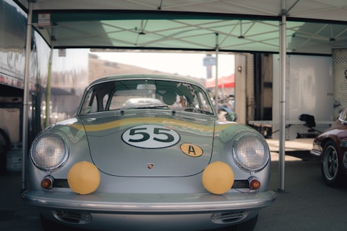 A vintage silver race car is parked under a canopy. The car features a racing number 55 on the hood and two yellow accents on the headlights. It is positioned in a setting typical for a motorsport event, with additional vehicles and equipment visible in the background.