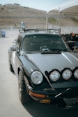 A vintage black sports car is parked outdoors under an open tent. The car has a sleek design with large round headlights and is fitted with additional driving lights on the front. It is equipped with off-road tires and a roof rack. In the background, there is a hilly landscape and a few scattered structures.