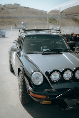 A vintage black sports car is parked outdoors under an open tent. The car has a sleek design with large round headlights and is fitted with additional driving lights on the front. It is equipped with off-road tires and a roof rack. In the background, there is a hilly landscape and a few scattered structures.