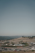 A racetrack is nestled within a hilly landscape, featuring winding roads and open spaces. White tents are scattered throughout the area, with some vehicles parked in the vicinity. The surrounding terrain is arid, offering a backdrop of mountains under a clear sky.