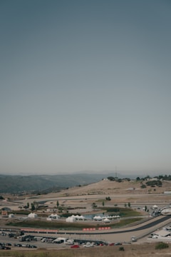 A racetrack is nestled within a hilly landscape, featuring winding roads and open spaces. White tents are scattered throughout the area, with some vehicles parked in the vicinity. The surrounding terrain is arid, offering a backdrop of mountains under a clear sky.