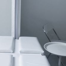 A minimalist indoor setting with a folded metal chair and a small round table positioned beside a cushioned bench near a corner. The wall and furnishings are in monochromatic shades, giving a clean and sterile look.