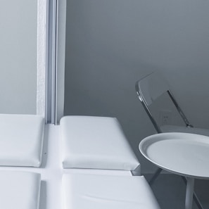 A minimalist indoor setting with a folded metal chair and a small round table positioned beside a cushioned bench near a corner. The wall and furnishings are in monochromatic shades, giving a clean and sterile look.