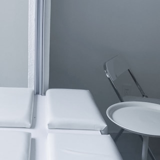 A minimalist indoor setting with a folded metal chair and a small round table positioned beside a cushioned bench near a corner. The wall and furnishings are in monochromatic shades, giving a clean and sterile look.