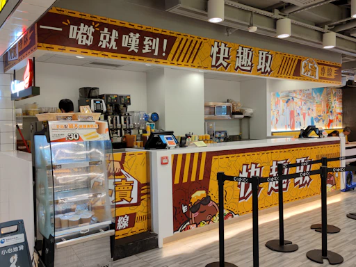 A fast-food kiosk with bright yellow and red signage displaying a variety of drinks and an electronic ordering system. Behind the counter, an assortment of beverage machines are present. The kiosk has a functional layout with a small queue area delineated by black stanchions. On the side, there is a vivid mural featuring cartoon-style illustrations.