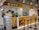 A fast-food kiosk with bright yellow and red signage displaying a variety of drinks and an electronic ordering system. Behind the counter, an assortment of beverage machines are present. The kiosk has a functional layout with a small queue area delineated by black stanchions. On the side, there is a vivid mural featuring cartoon-style illustrations.