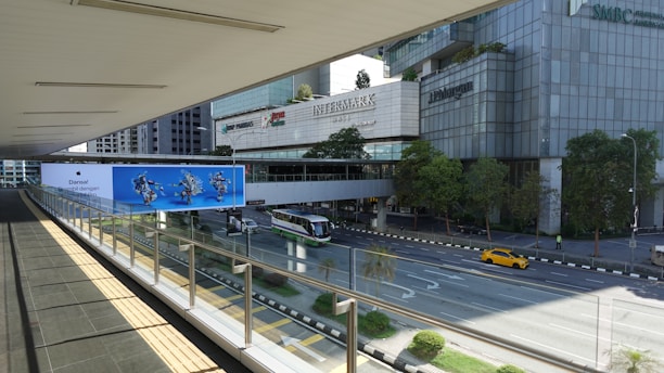 A pedestrian bridge with a glass railing overlooks a multi-lane road. Below, there's a green and white city bus and a yellow car on the road. On the opposite side, there are tall office buildings with logos including 'BNP Paribas', 'Intermark', and 'J.P. Morgan'. Trees line the sidewalk, and there is a large blue advertisement featuring images of people in motion.