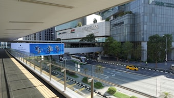 A pedestrian bridge with a glass railing overlooks a multi-lane road. Below, there's a green and white city bus and a yellow car on the road. On the opposite side, there are tall office buildings with logos including 'BNP Paribas', 'Intermark', and 'J.P. Morgan'. Trees line the sidewalk, and there is a large blue advertisement featuring images of people in motion.