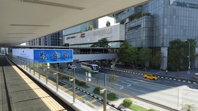 A pedestrian bridge with a glass railing overlooks a multi-lane road. Below, there's a green and white city bus and a yellow car on the road. On the opposite side, there are tall office buildings with logos including 'BNP Paribas', 'Intermark', and 'J.P. Morgan'. Trees line the sidewalk, and there is a large blue advertisement featuring images of people in motion.